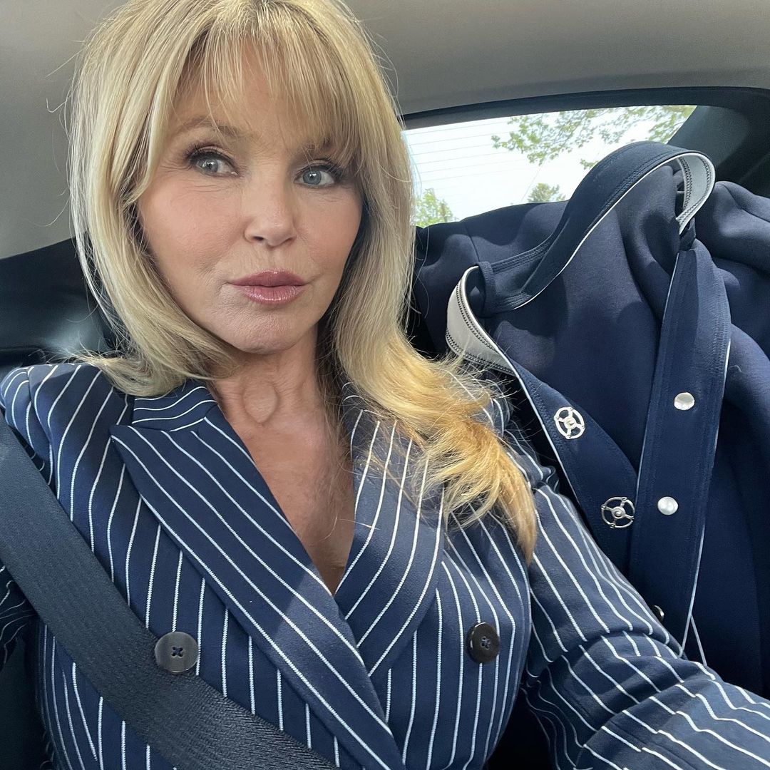 Christie Brinkley in a car, wearing a striped blue blazer with a seatbelt across her chest.