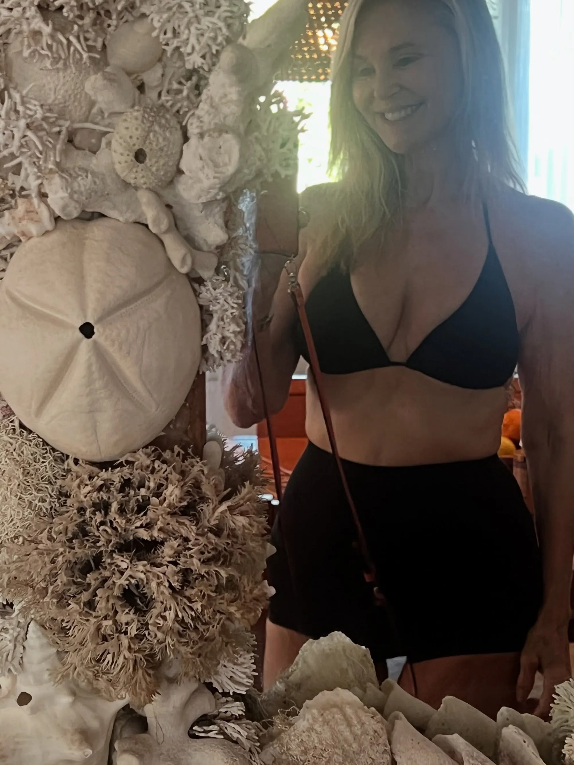 Christie Brinkley wearing a black bikini top and black shorts, smiling next to a column adorned with shells and corals.