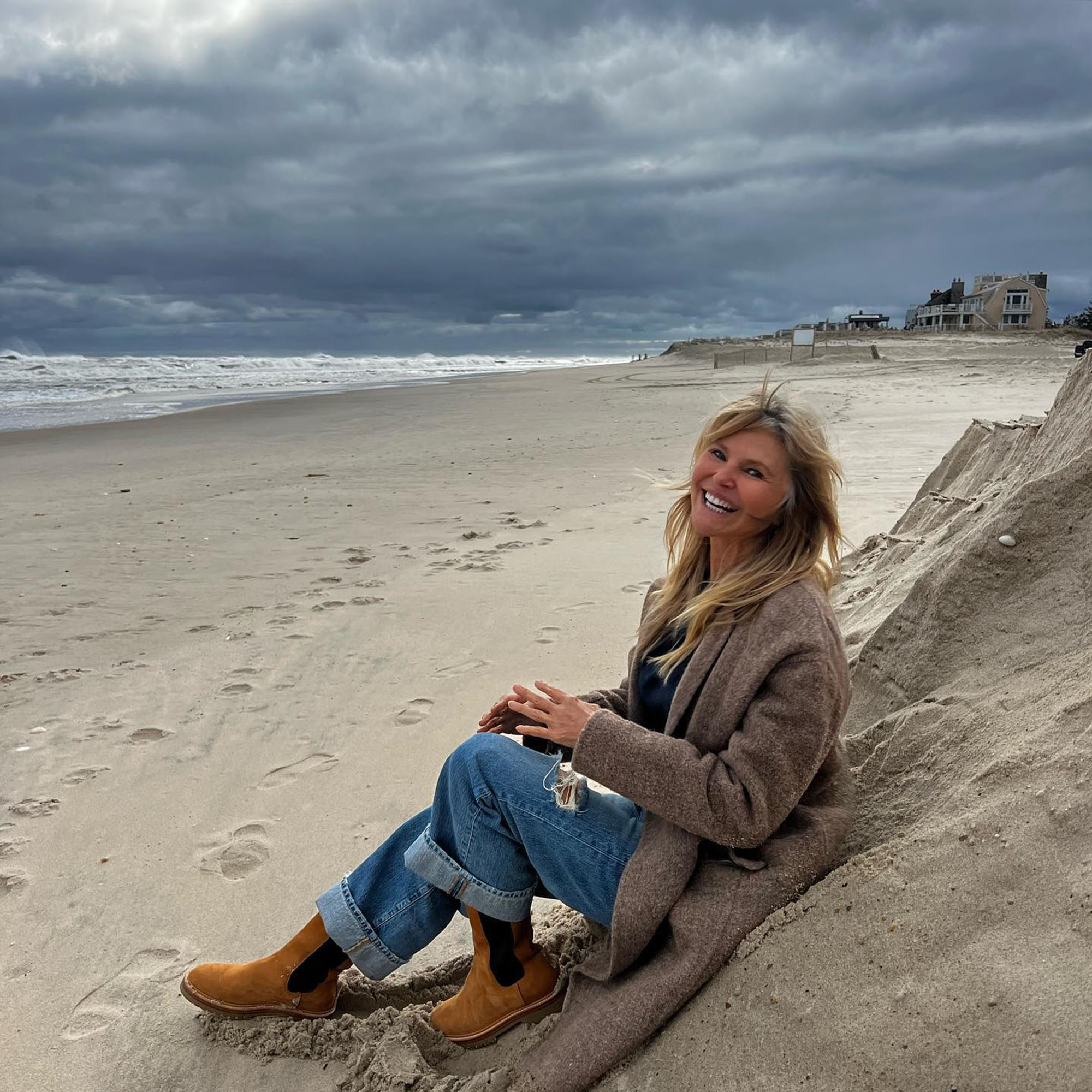 Christie Brinkley sits on a sandy beach, smiling at the camera, with the ocean and storm clouds in the background.