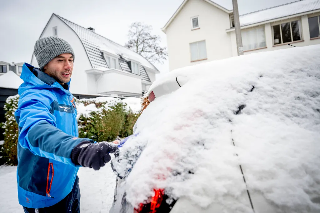 A man in a blue jacket and grey beanie clearing snow from a car in Utrecht.