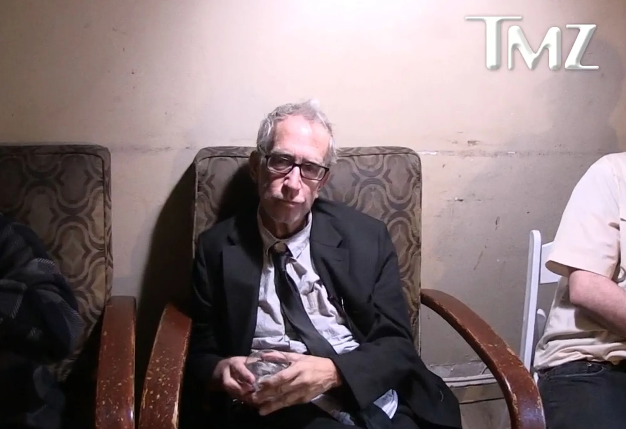 Andy Dick sitting in a chair, wearing glasses and a suit, holding a glass.
