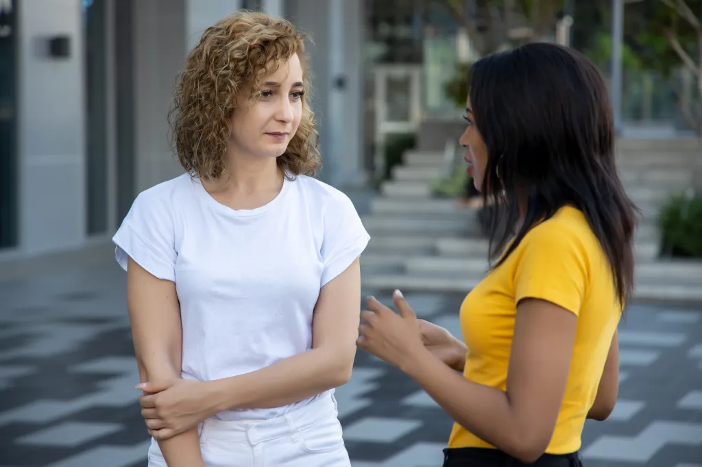 Two women, one with blonde curly hair and the other with dark straight hair, talk with each other.