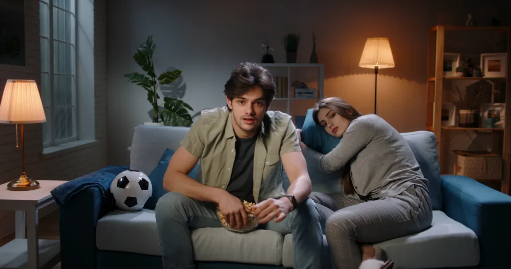 A man animatedly watches football on a couch with popcorn while a woman next to him appears bored and lies her head on a cushion.