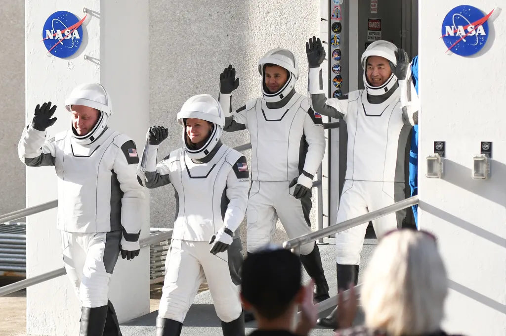 Astronauts Mike Fincke, Zena Cardman, Oleg Platonov, and Kimiya Yui in white suits and helmets wave to onlookers.