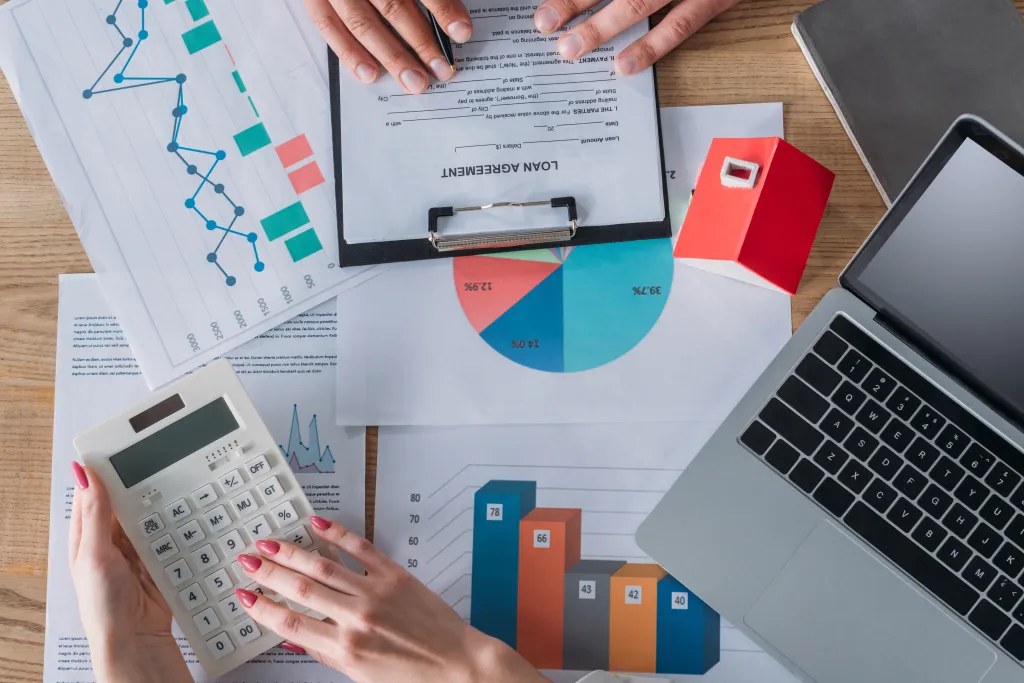 Overhead shot of a man reviewing a loan agreement and a woman using a calculator at a desk with a laptop, house model, graphs, and charts.