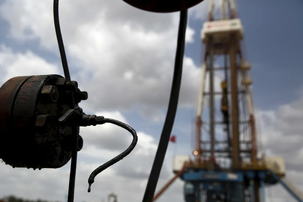 Crude oil drips from a valve at an oil well, with a drilling rig in the background.
