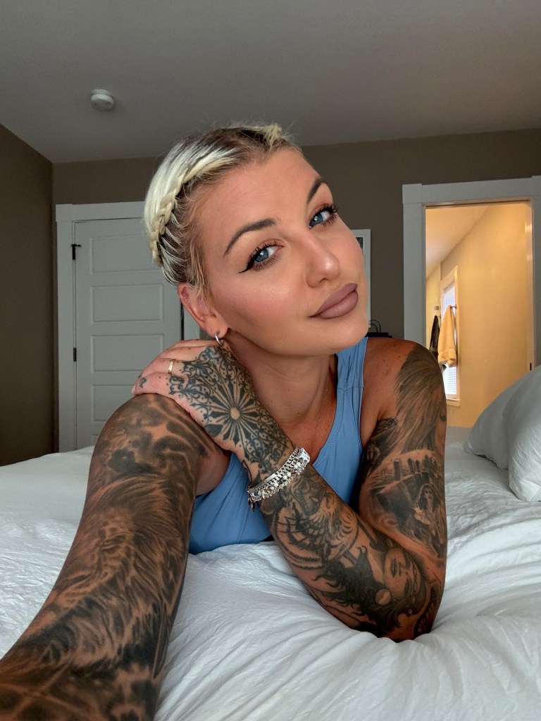 A woman with braided blonde hair and heavily tattooed arms lies on a white bed, looking at the camera.
