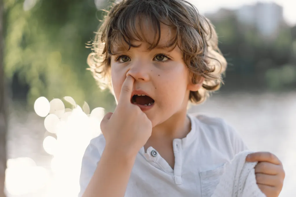 A young boy with curly hair picking his nose, with his mouth open, outdoors.