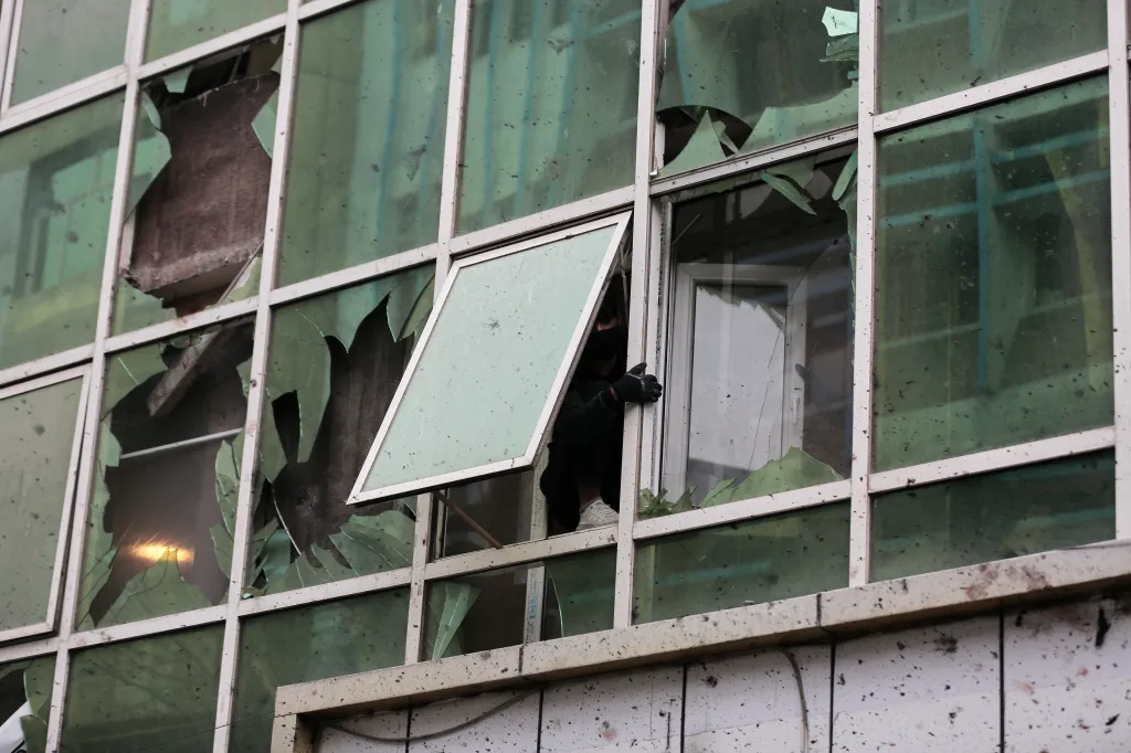 A person in a black mask and gloves stands in a broken window of a damaged building.