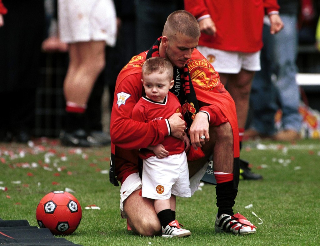 David Beckham, in a red England soccer strip, holding his son Brooklyn, in a red football jersey and white shorts, after a Manchester United football match.