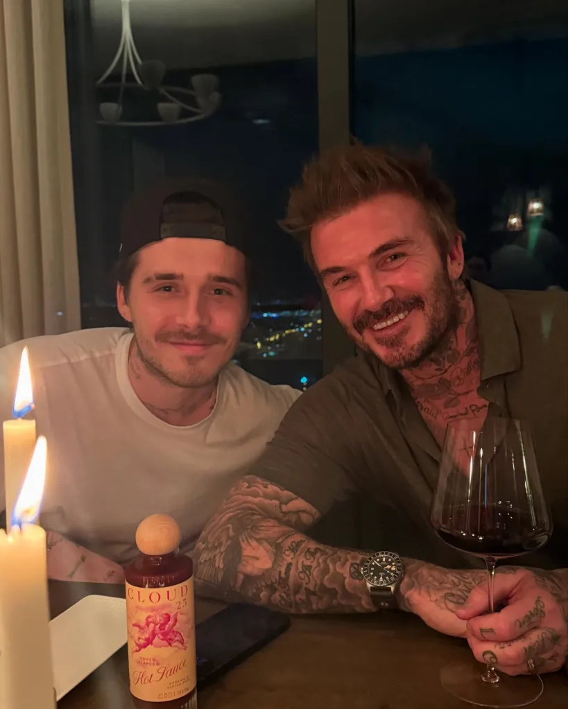 David and Brooklyn Beckham smiling at the camera at a table lit by candles, with a bottle of hot sauce and a glass of red wine visible.