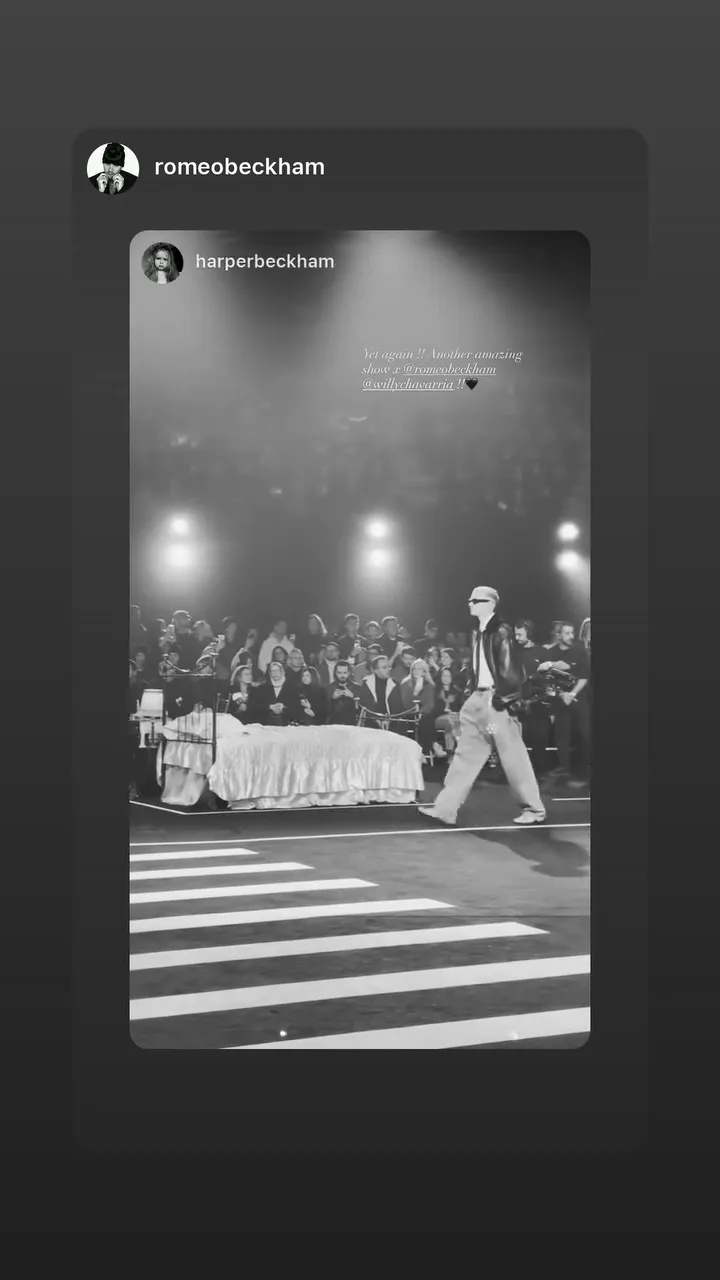Romeo Beckham modeling in an all-black fashion show, walking past a bed on a runway with an audience.