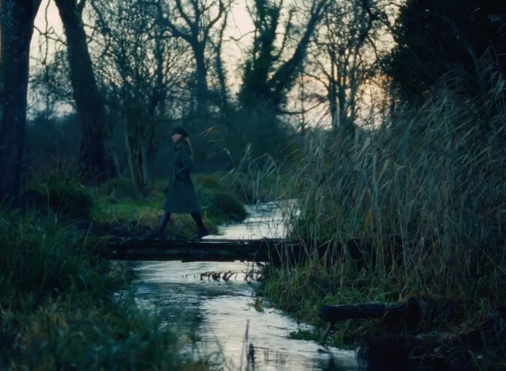 A person in a long coat crossing a stream on a small wooden bridge.