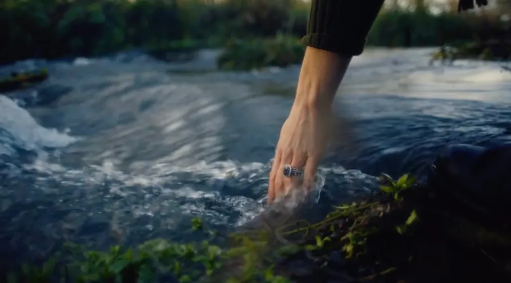 A person's hand with a ring touching the flowing water of a river.