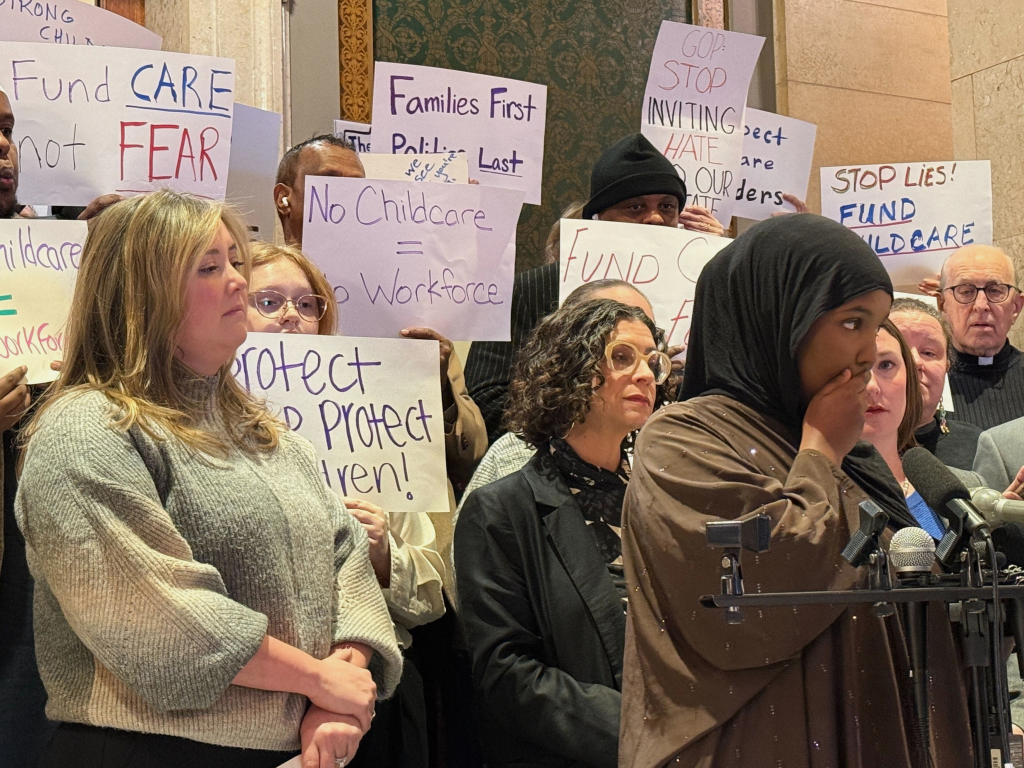 Deko Nor speaking at a news conference in St. Paul, Minnesota, surrounded by people holding signs about childcare funding and politics.