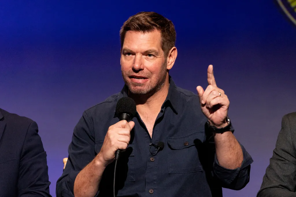 Eric Swalwell speaks into a microphone, gesturing with his right hand, in front of a blue background.