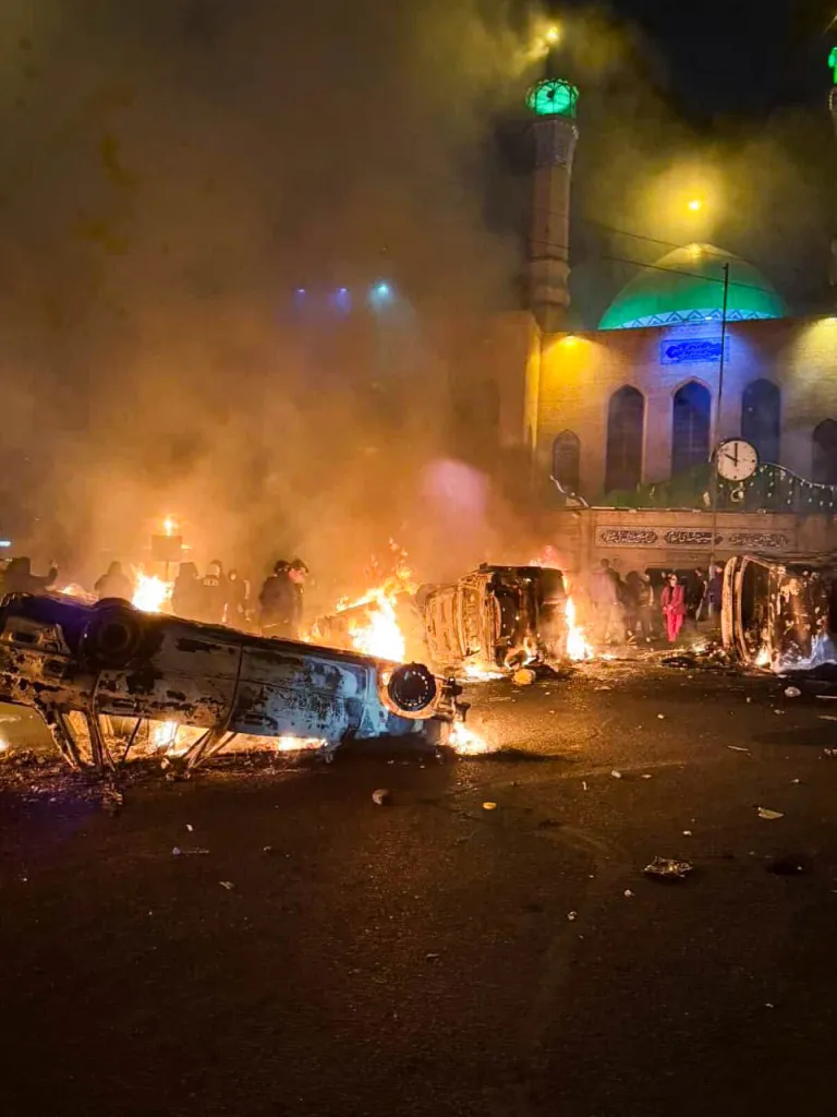 Burnt cars and debris litter a street in front of a mosque at night during protests in Tehran.