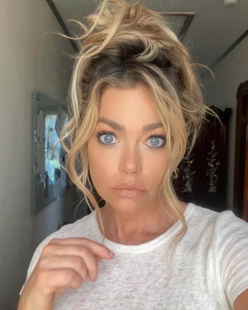 Denise Richards looks directly at the camera with wide blue eyes, wearing a white t-shirt and her blonde hair in a messy bun.