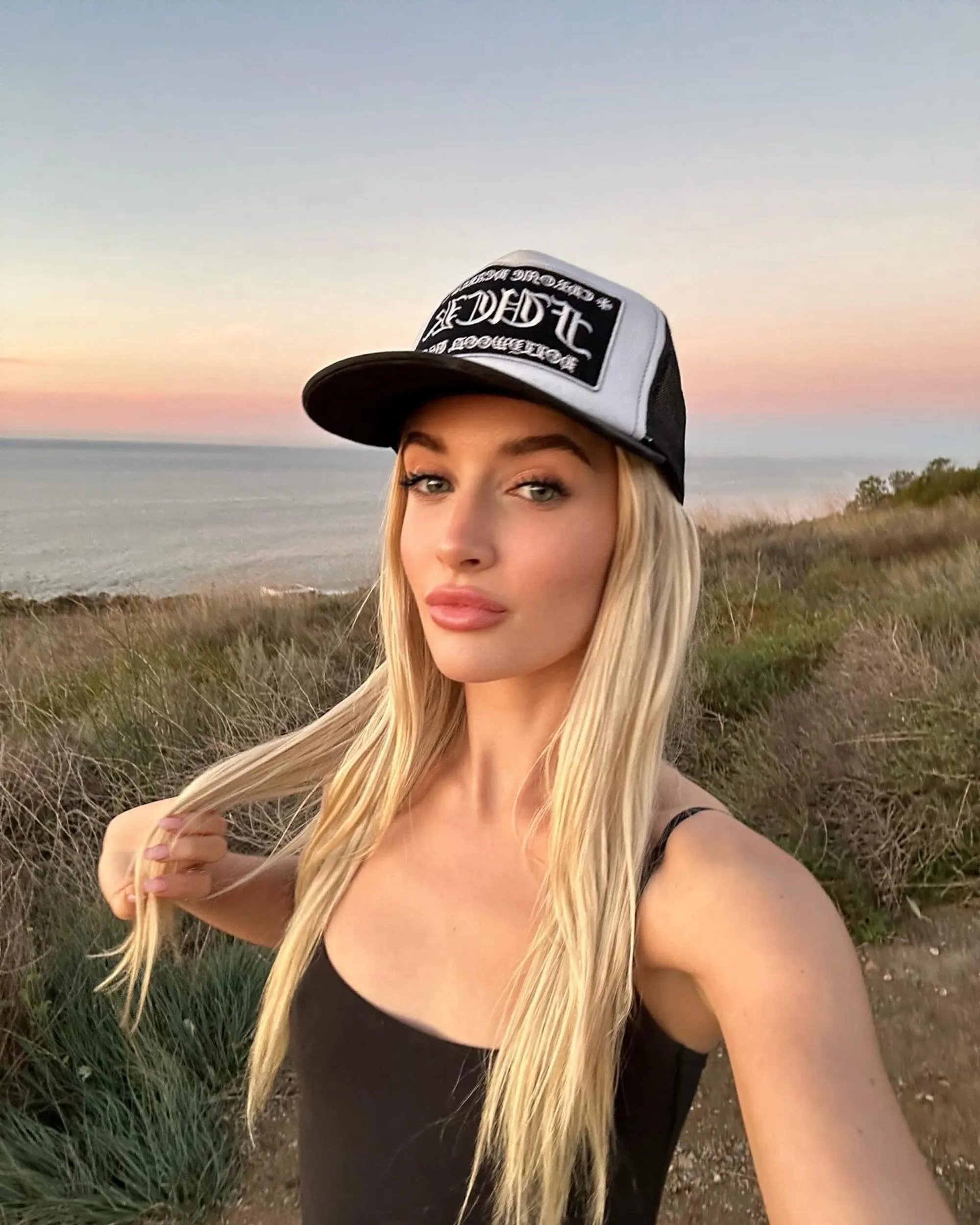 Sami Sheen wearing a trucker hat and black top, holding her long blonde hair, with an ocean sunset in the background.