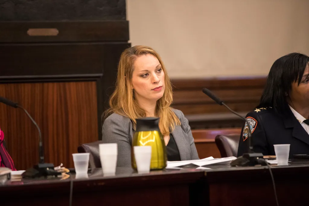 A woman with long blonde hair and a gray blazer sits at a long table with a microphone, paper cups, and a yellow thermos.