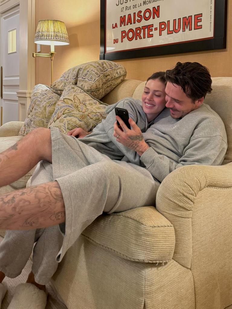 Brooklyn Beckham, in a grey sweatshirt and grey shorts, and Nicola Peltz, in a grey sweatshirt and grey sweatpants, looking at a phone while sitting on a couch.