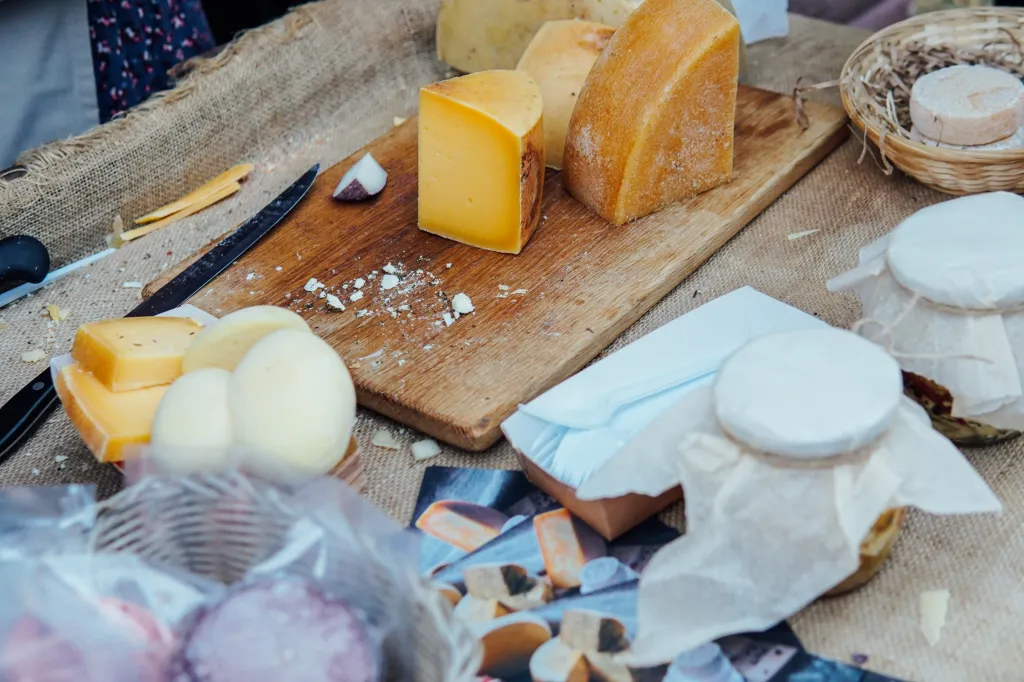 Various types of cheese on a rustic table, including wedges on a wooden board and smaller rounds wrapped in paper.
