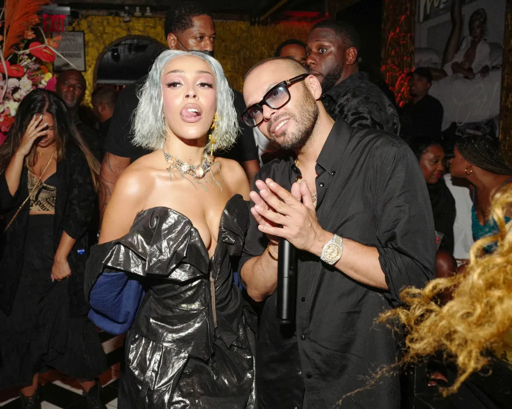 Doja Cat in a metallic off-the-shoulder dress sticking out her tongue next to Richie Akiva in a black button-down shirt and sunglasses.