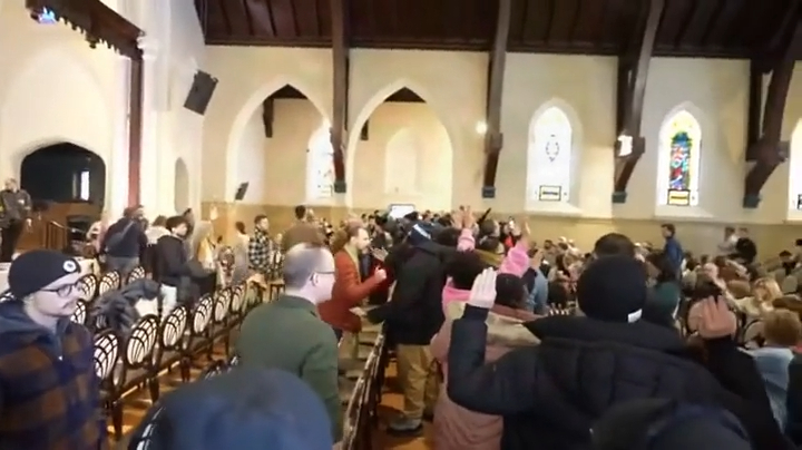 People gathered in a church, some standing and some sitting in rows of chairs, with gothic arches and stained-glass windows visible.