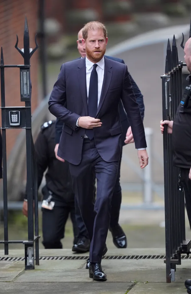 Prince Harry, Duke of Sussex, walking through a gate while wearing a dark suit and tie.