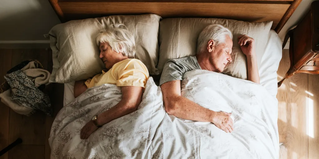 An elderly couple, a woman in a yellow shirt and a man in a gray shirt, sleeping peacefully in bed