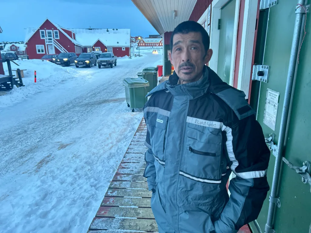 Elias Lunge, a Greenland resident, stands outdoors in winter attire with snow-covered buildings and cars in the background.