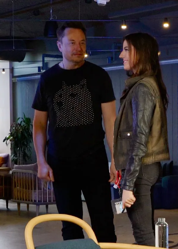 Elon Musk in a black shirt speaks with a woman in a brown and black jacket.
