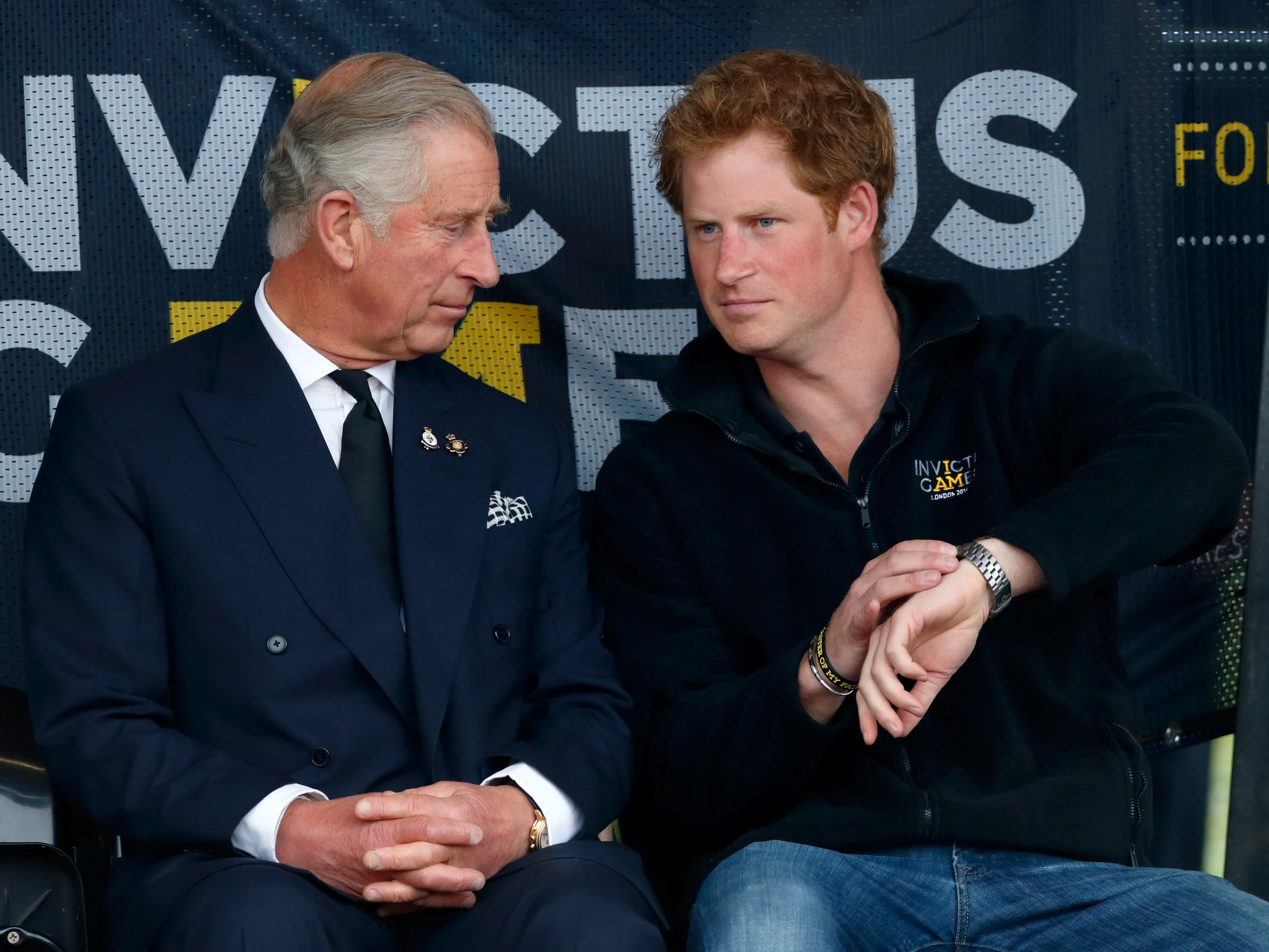 Prince Charles and Prince Harry sit together, looking at each other, against a banner for the Invictus Games.