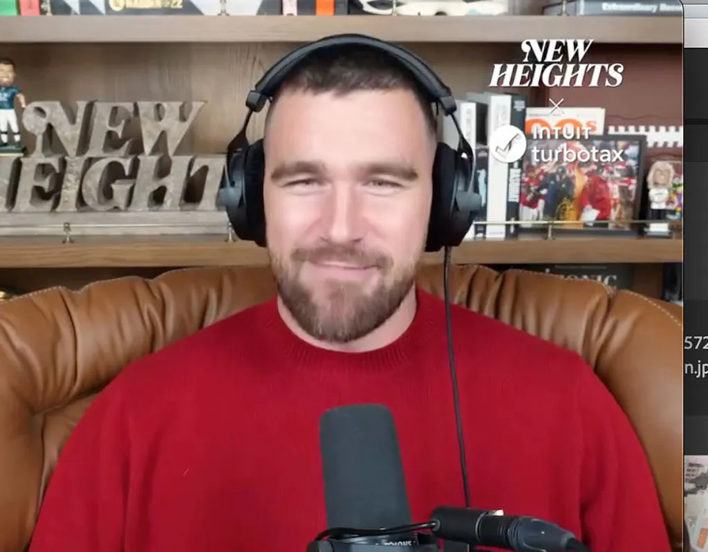 Travis Kelce wearing a red sweater and headphones, sitting in a brown leather chair in front of a “New Heights” sign.