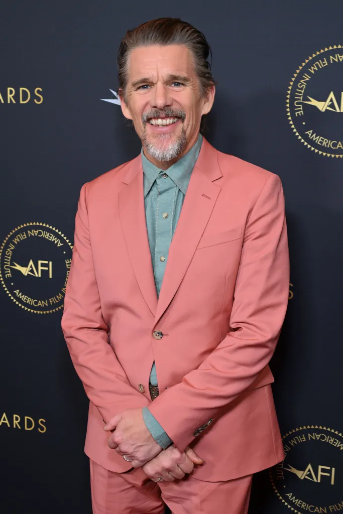 Ethan Hawke smiles, wearing a coral suit over a light blue collared shirt, at the AFI Awards.
