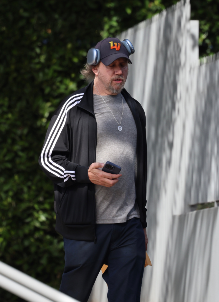 Jamie Kennedy in an Adidas jacket, jeans, and Skechers sneakers, wearing a cap with headphones, holding a phone, on his way to the bank.