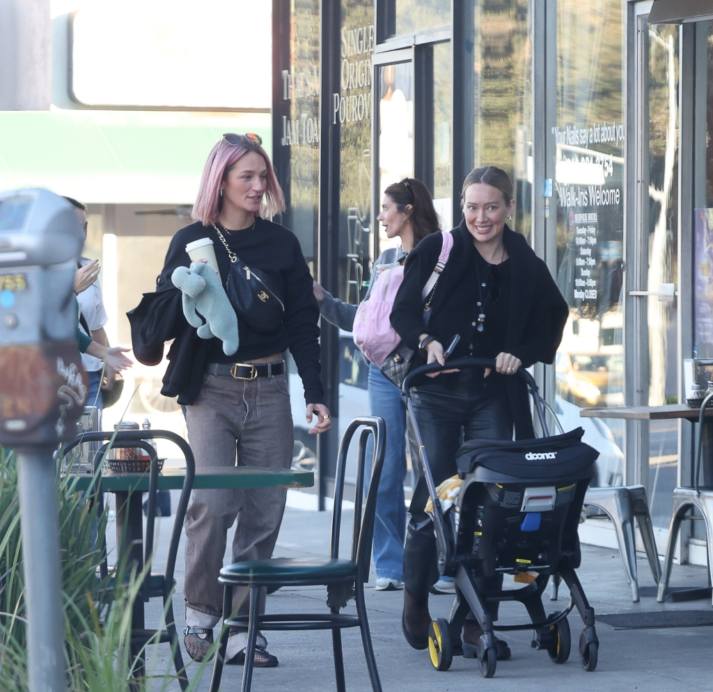 Hilary Duff pushing a Doona baby stroller next to a friend with pink hair, both smiling, walking past outdoor cafe seating.