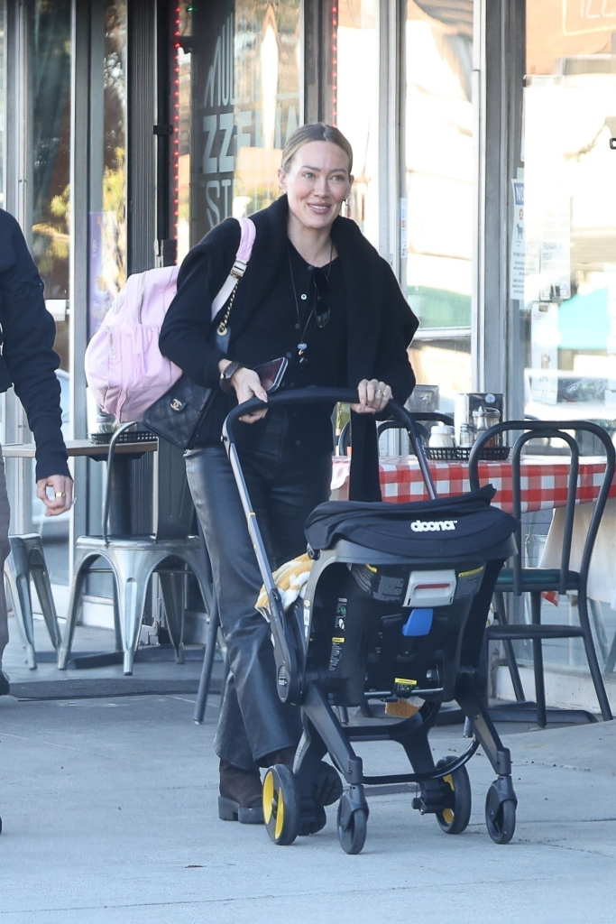 Hilary Duff smiles while pushing a baby stroller outdoors.