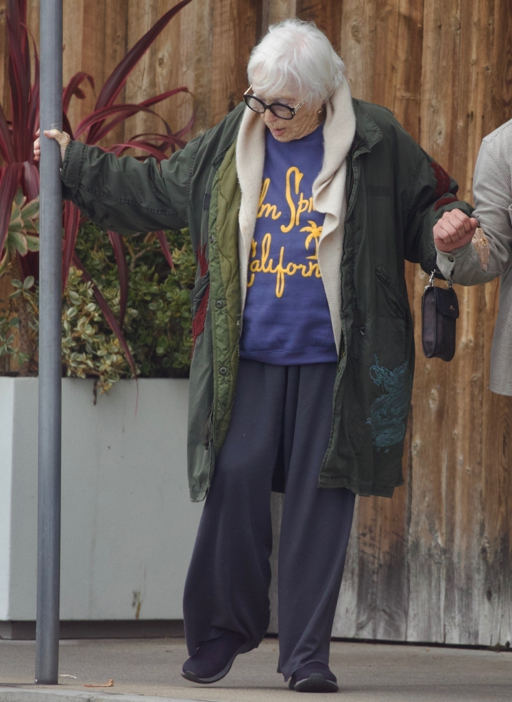Shirley MacLaine in a dark green coat, blue sweatshirt, and dark sweatpants holding onto a pole.