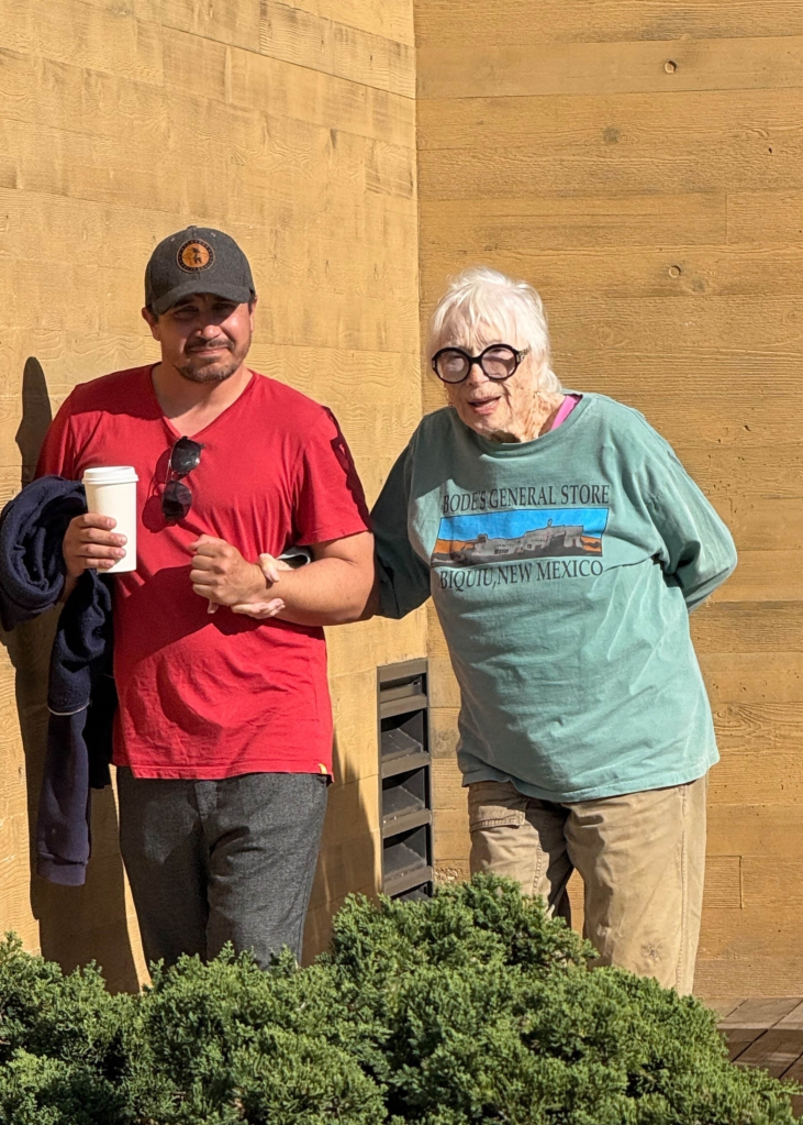 Shirley MacLaine with a man assisting her, standing in front of a textured wall.