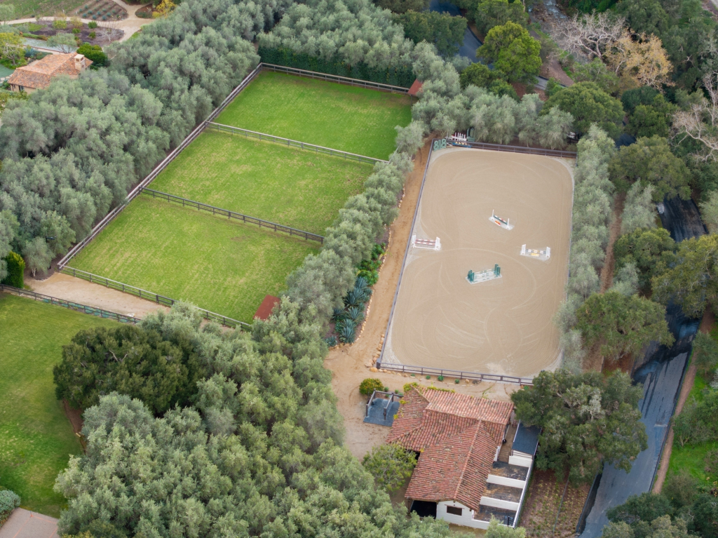 Aerial view of Kendall Jenner's Montecito estate with a horse stable, riding arena with show jumping facilities, and adjacent green pastures, surrounded by trees.
