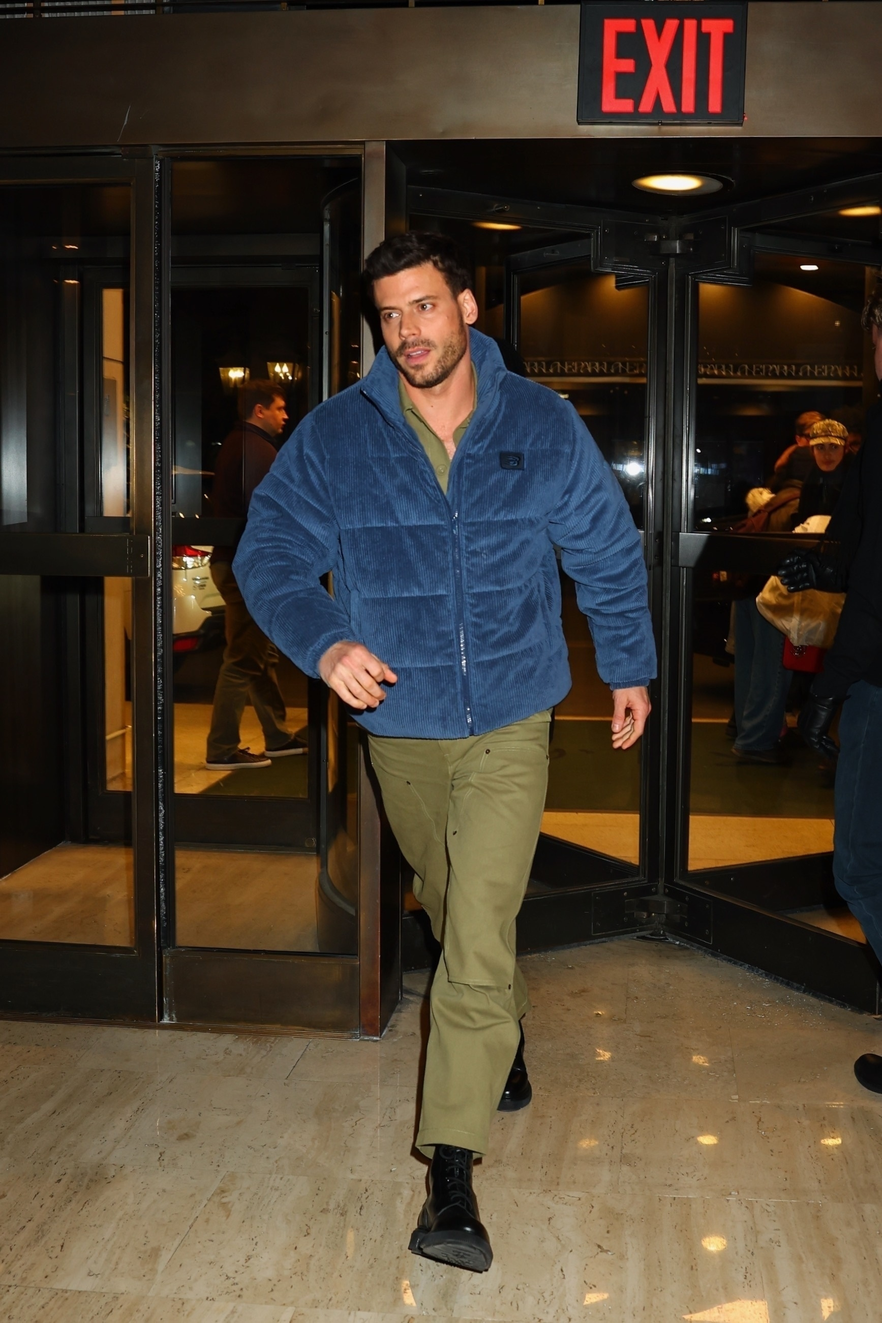 Francois Arnaud in a blue jacket and olive pants walking out of a building.