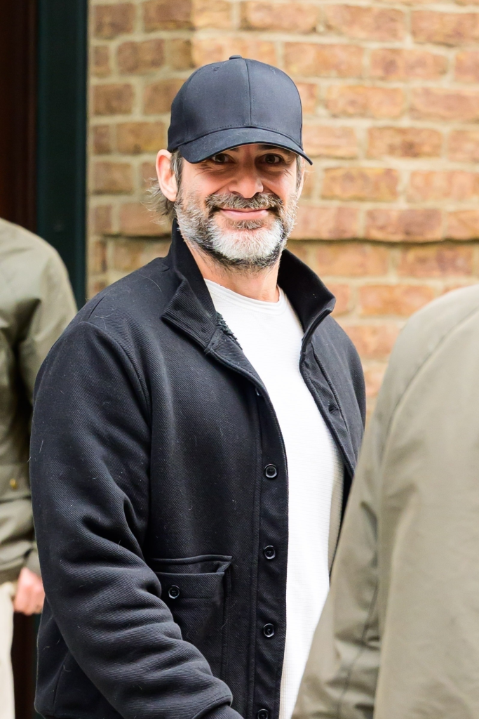 Jim Curtis, Jennifer Aniston's boyfriend, smiling in a black bomber jacket, white shirt, and baseball cap in NYC.