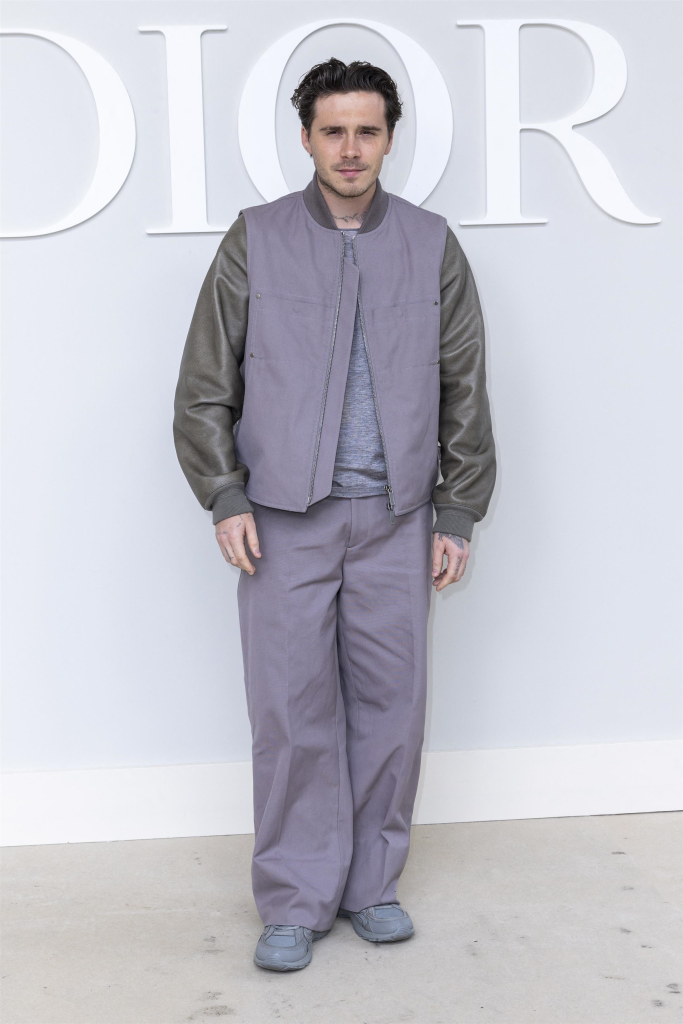 Brooklyn Peltz Beckham wearing a gray bomber jacket with dark green leather sleeves and matching gray trousers, standing in front of a white wall with the partial letters 