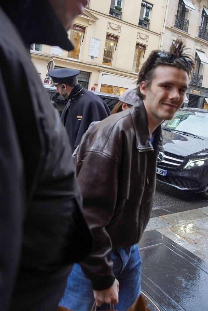 Cruz Beckham in a brown leather jacket and blue jeans, carrying a bag, walks on a wet street.