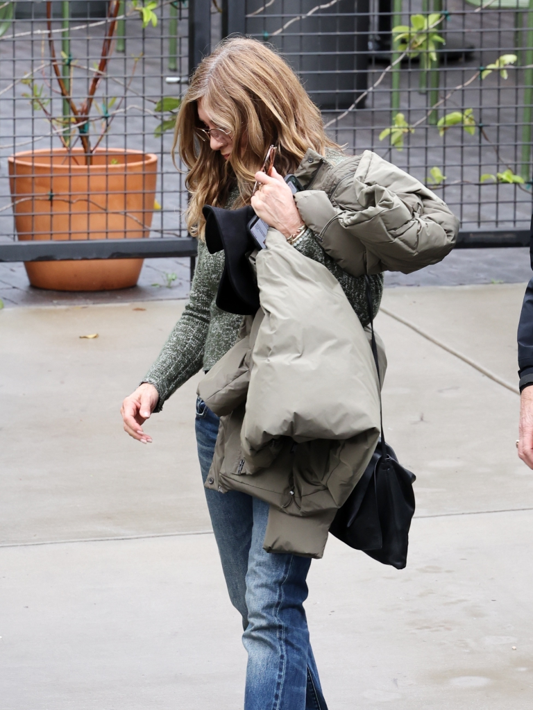Jennifer Aniston holding a jacket and bag, wearing blue jeans and a green sweater.