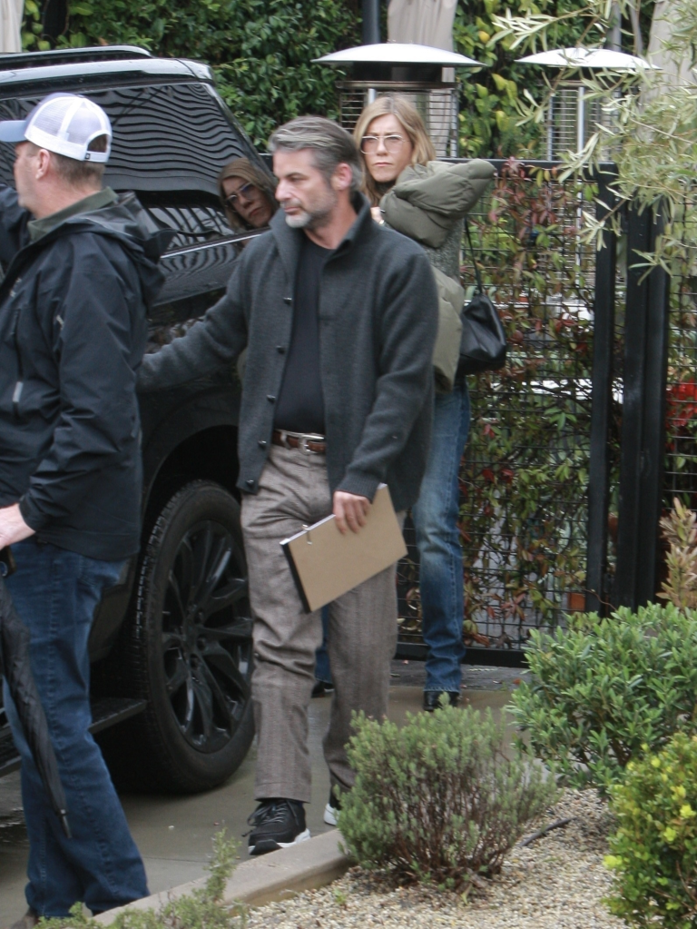 Jennifer Aniston looking over the shoulder of Jim Curtis, as he walks carrying a clipboard near a black car.