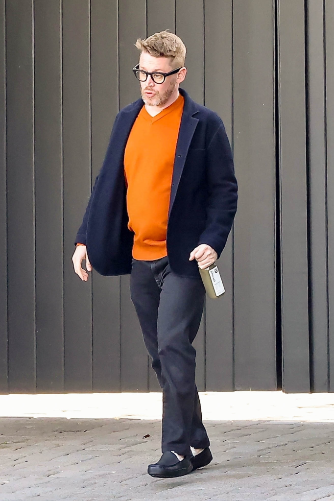 Macaulay Culkin wearing an orange sweater, blue blazer, black pants, glasses, and a beard, walking on a paved surface.