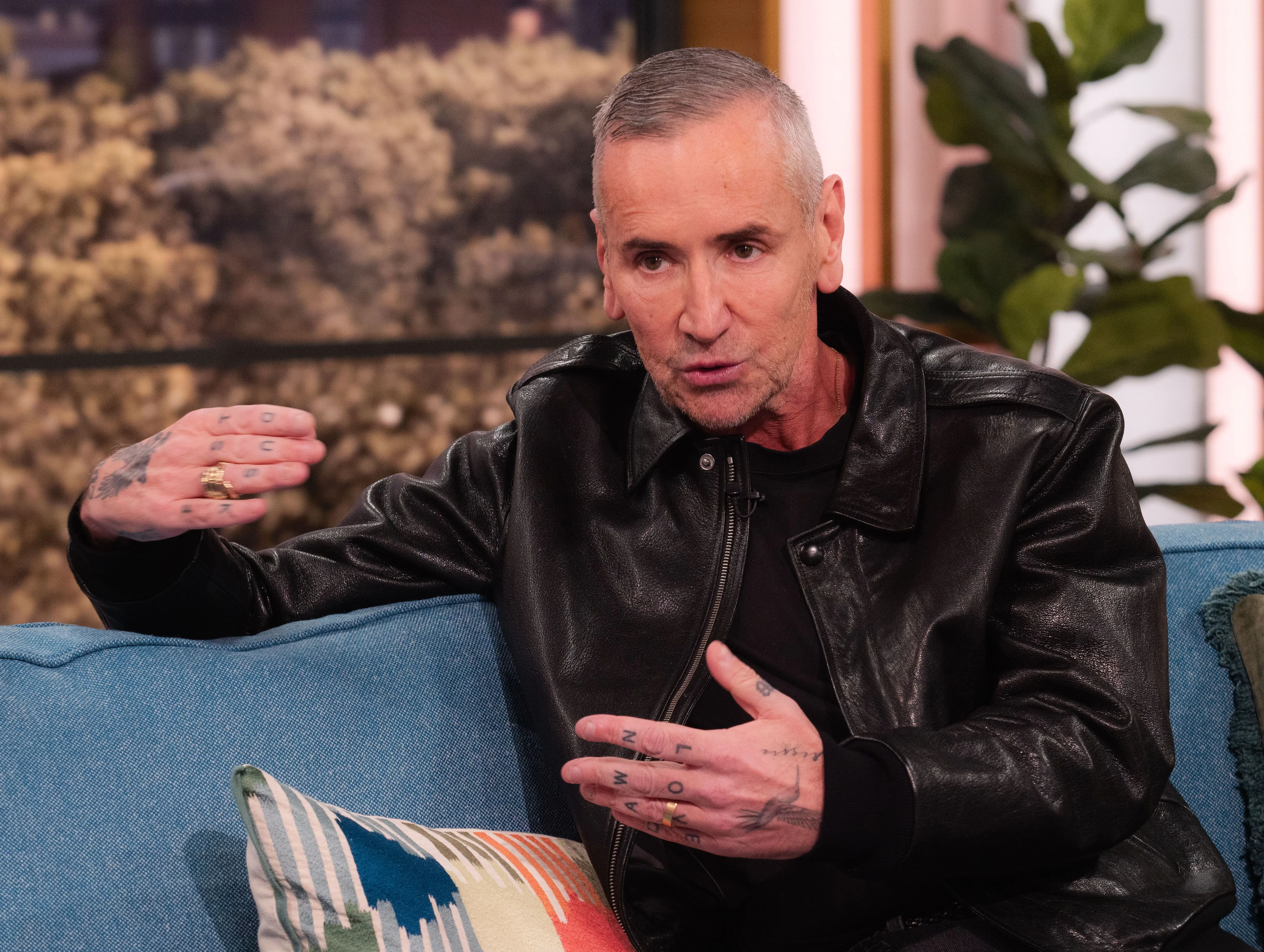 DJ Fat Tony wearing a black leather jacket, sitting on a blue sofa, and gesturing with his tattooed hand during an interview on 