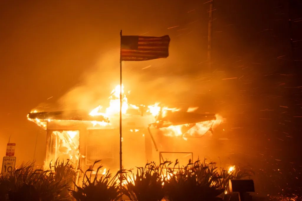 A house engulfed in flames during the Palisades Fire, with an American flag flying on a pole in front.